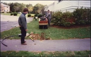 Two crew members installing pipe with a trenching machine along a sidewalk